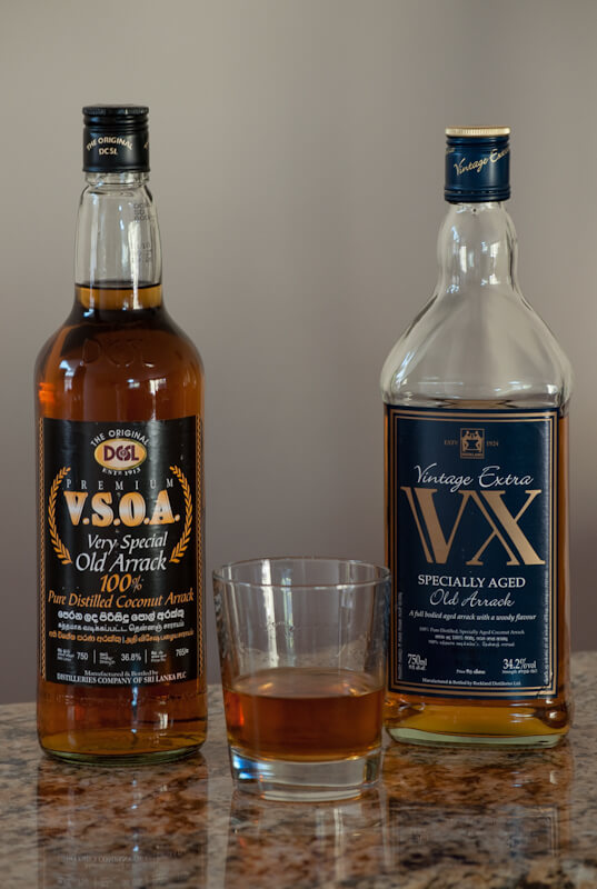 兩瓶亞拉克酒，V.S.O.A 和 VX，擺放在大理石表面。中間放有一杯亞拉克酒，展現出琥珀色的酒體。