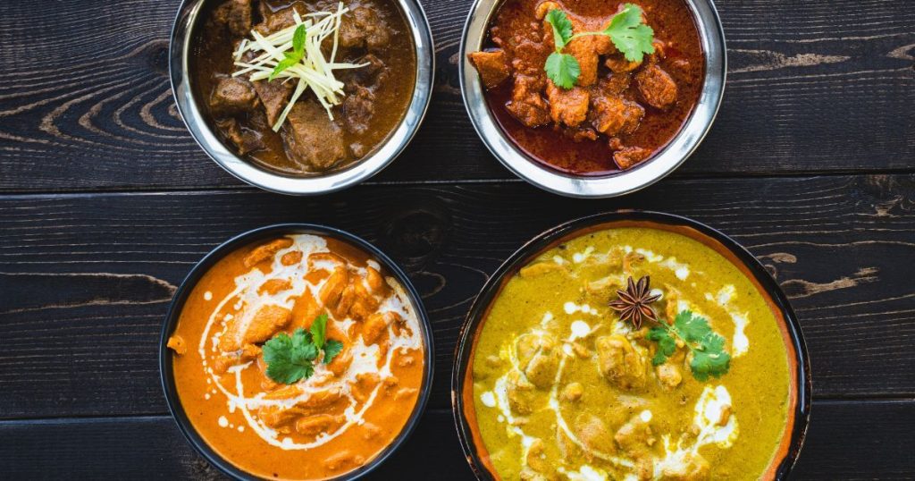 四碗色彩鮮豔的咖哩，從紅色到橙色再到綠色，餐盤上裝飾著香草，擺放在深色木質餐桌上。