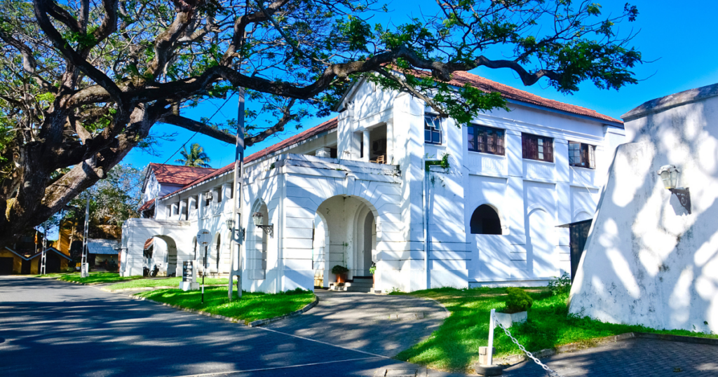 圖片展示位於斯里蘭卡加耳(Galle)的白色殖民建築——法院大樓。建築結構古典優雅,屋頂覆蓋紅瓦,周圍有翠綠的草坪和高大的樹木,陽光穿透枝葉形成美麗的光影效果,反映斯里蘭卡殖民時期的歷史建築風格與當地的熱帶自然風情。
