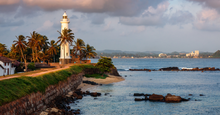 這張圖片展示了斯里蘭卡著名的加勒燈塔（Galle Lighthouse），燈塔周圍棕櫚樹環繞，位於海岸邊的石牆上，與藍色的大海形成對比。背景遠處可以看到城鎮建築，天際線呈現日落時分的柔和色調，傳遞出悠閒與和平的氣氛。