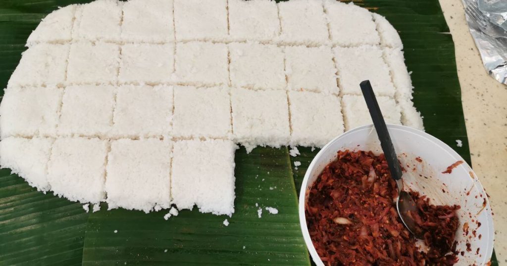 這張照片展示了切成小方塊的Kiribath（椰漿飯），旁邊擺放了一碗伴菜，背景為傳統的蕉葉餐桌佈置。