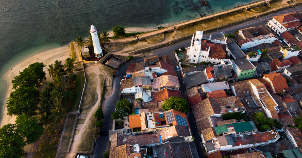 圖片展示了斯里蘭卡著名的加勒古堡(Galle Fort)及燈塔的全景鳥瞰圖。燈塔位於海岸邊,周圍環繞著熱帶椰子樹和白色沙灘,與碧藍的大海形成鮮明對比。畫面中的古堡區域充滿殖民時期的建築風格,紅瓦屋頂與整齊排列的街道增添了歷史氛圍。這張圖片不僅展現了加勒古堡作為世界文化遺產的獨特魅力,也反映了斯里蘭卡濃厚的歷史與自然風光的交融。