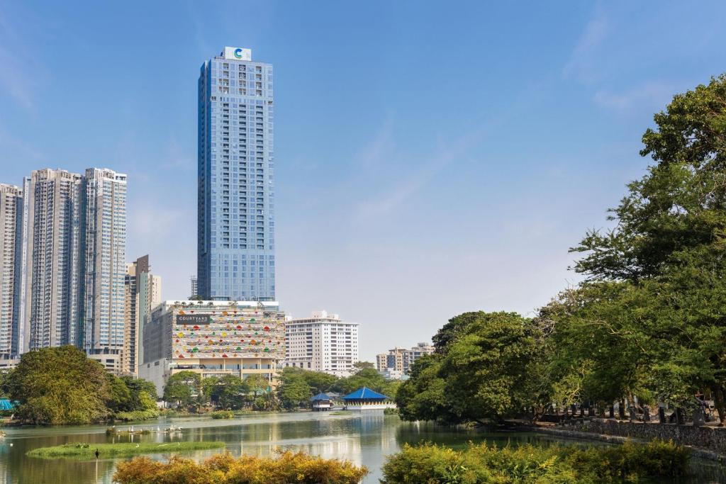這張圖片展示了Courtyard by Marriott Colombo飯店位於都市中心的高樓外觀，建築外觀高聳且現代，周圍是林立的建築和湖泊，城市天際線與自然景色相互交融，呈現出一幅美麗的城市景觀。