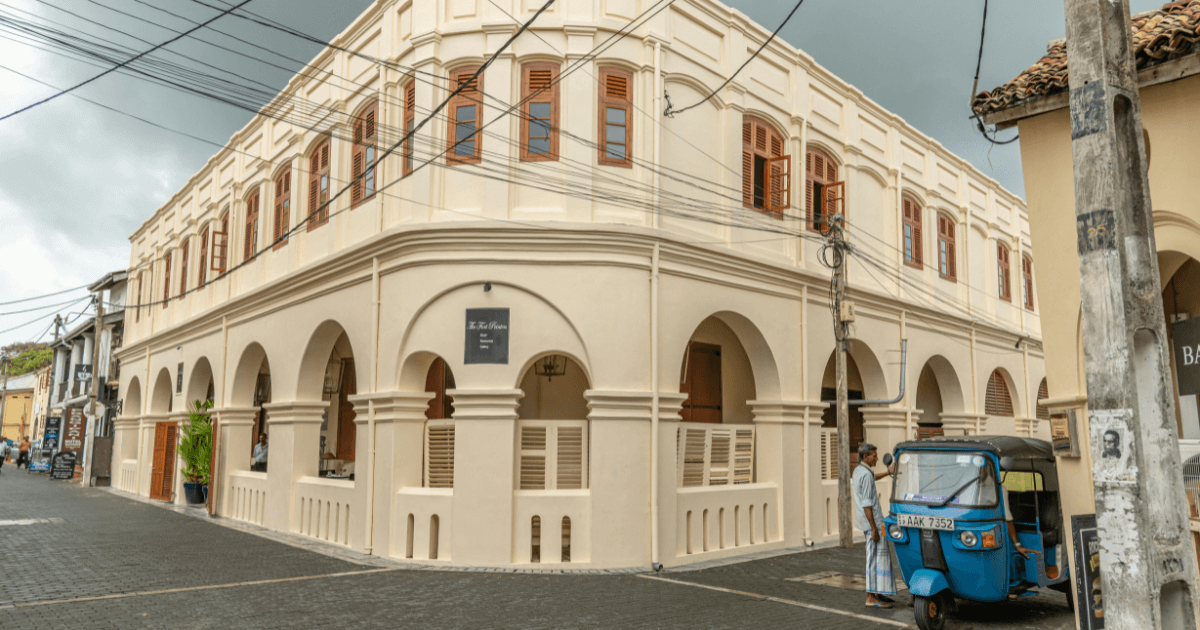 這張圖片展示了斯里蘭卡加勒（Galle）的街道，畫面中可見一輛藍色的三輪車（嘟嘟車）停在街角。旁邊是經典的殖民風格建築，展現出當地獨特的歷史與文化氛圍。街道上有一些當地人和商店，整體氛圍輕鬆又富有生活氣息，這是加勒古城的典型景象。