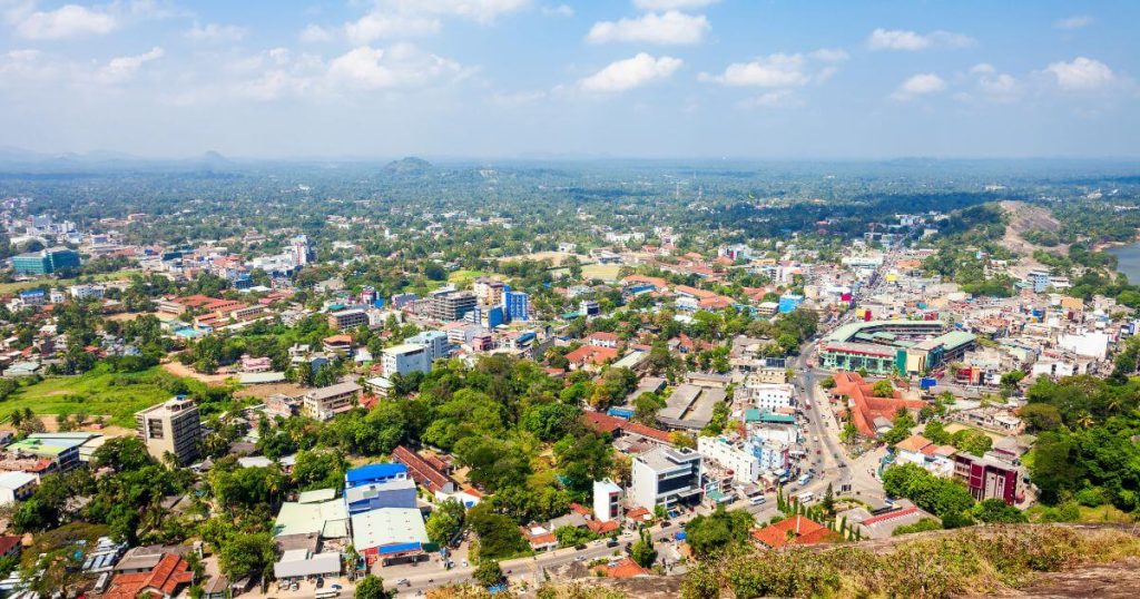 此圖片展示了斯里蘭卡庫魯內格勒市的全景,俯瞰整個城市,能看到市中心的各式建築及廣大的綠地。藍天白雲之下,遠處的山巒和河流隱約可見,反映出這個城市與自然環境的融合與和諧。