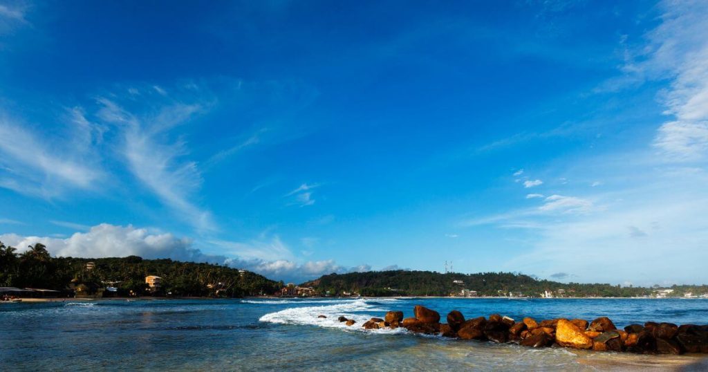 這張圖片展示了斯里蘭卡米瑞莎海灘的風景，天空碧藍，海浪輕輕拍打沙灘，周圍環繞著翠綠的樹木和山丘，展現了寧靜的海邊度假勝地。