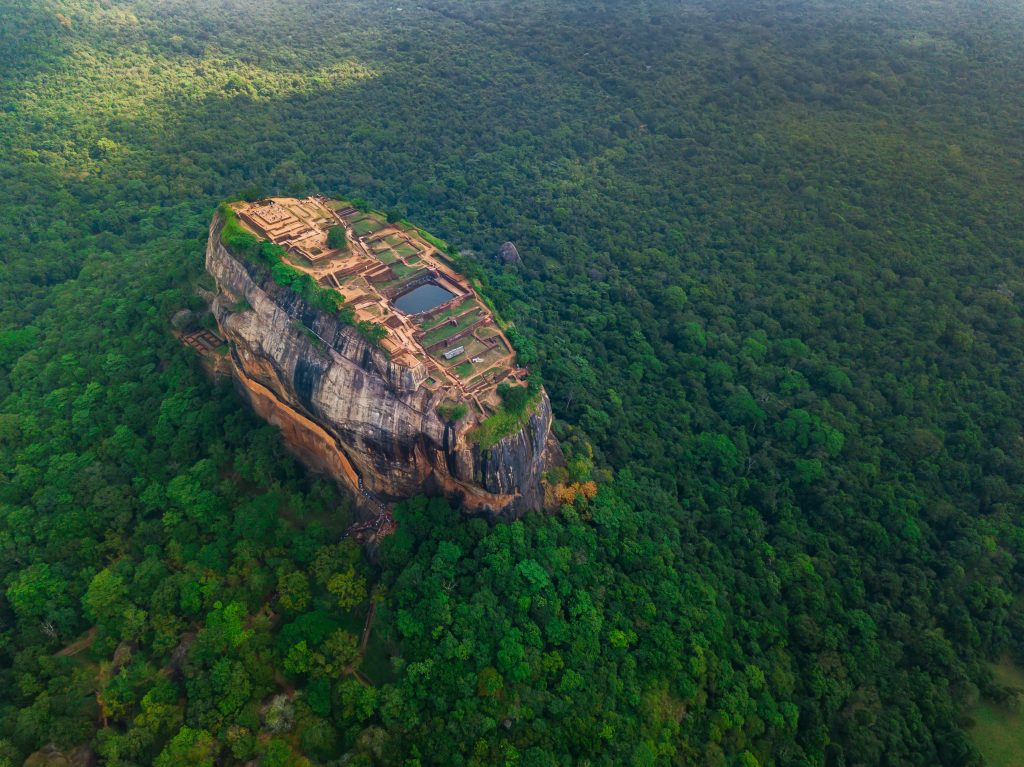 該圖片展示了斯里蘭卡著名的獅子岩(Sigiriya Rock)在青翠茂密的叢林環繞中,壯麗的古代石頭建築遺址從高處俯瞰,突顯出自然與歷史的和諧共存。
