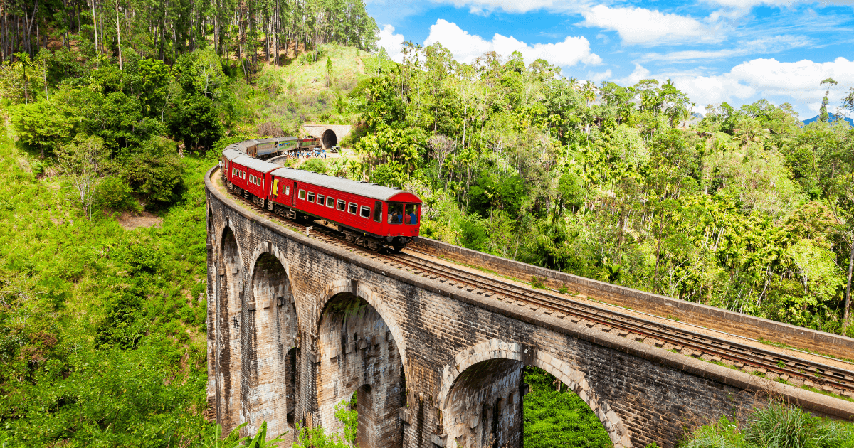 這張照片拍攝於斯里蘭卡（Sri Lanka）著名的九拱橋（Nine Arch Bridge），畫面中的紅色列車正行駛於古老的石橋之上，橋身拱形優美，位於綠意盎然的叢林與山林環繞中。天氣晴朗，藍天白雲搭配鮮紅列車構成強烈對比，展現出斯里蘭卡經典的慢旅風情與高原鐵道魅力。