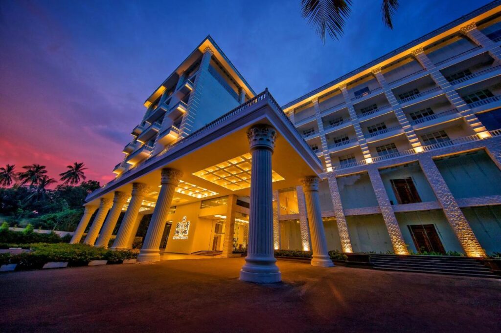 這張照片展現斯里蘭卡康提(Kandy)的 The Golden Crown Hotel 在夜色下的壯麗外觀。建築採用古典柱廊設計,燈光從底部照射至整棟樓層,突顯出典雅華麗的風格。夕陽映照的天空與金色光影交織,營造出夢幻的迎賓氛圍,是朝聖旅途中舒適入住的高級選擇之一。