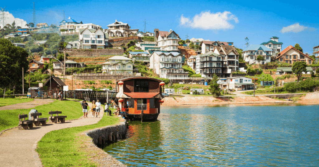 圖片展現斯里蘭卡努瓦勒埃利耶(Nuwara Eliya)湖畔的恬靜景色,湖水碧綠映照著藍天白雲,岸邊停泊著一艘紅色木船,吸引遊客駐足拍照。小鎮沿坡而建,整齊排列著帶有歐風建築特色的民宅,屋頂色彩多樣,綠意點綴其間。湖邊的步道與長椅增添休閒氛圍,整體呈現悠閒愜意的度假情境。
