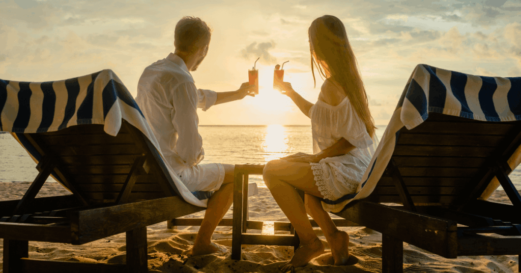 一對情侶坐在海邊的躺椅上,手中舉著果汁對飲,夕陽灑在海面上閃耀金光,氣氛浪漫而寧靜。畫面象徵蜜月旅行中專屬的親密時刻,展現兩人共享美好時光的幸福氛圍。