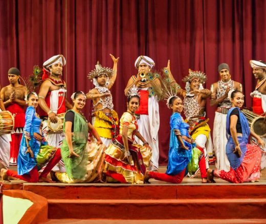 這張圖片展示了一群穿著色彩鮮豔傳統服飾的舞者，進行斯里蘭卡康提舞蹈表演。舞者們的動作充滿力量和優雅，身穿紅、藍、綠等色彩的服裝，搭配各種傳統樂器，包括大鼓和小鼓，背景是一道深紅色的舞台布幕。