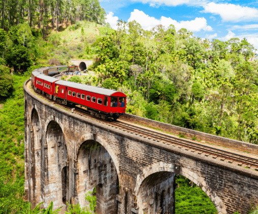 這張照片拍攝於斯里蘭卡（Sri Lanka）著名的九拱橋（Nine Arch Bridge），畫面中的紅色列車正行駛於古老的石橋之上，橋身拱形優美，位於綠意盎然的叢林與山林環繞中。天氣晴朗，藍天白雲搭配鮮紅列車構成強烈對比，展現出斯里蘭卡經典的慢旅風情與高原鐵道魅力。
